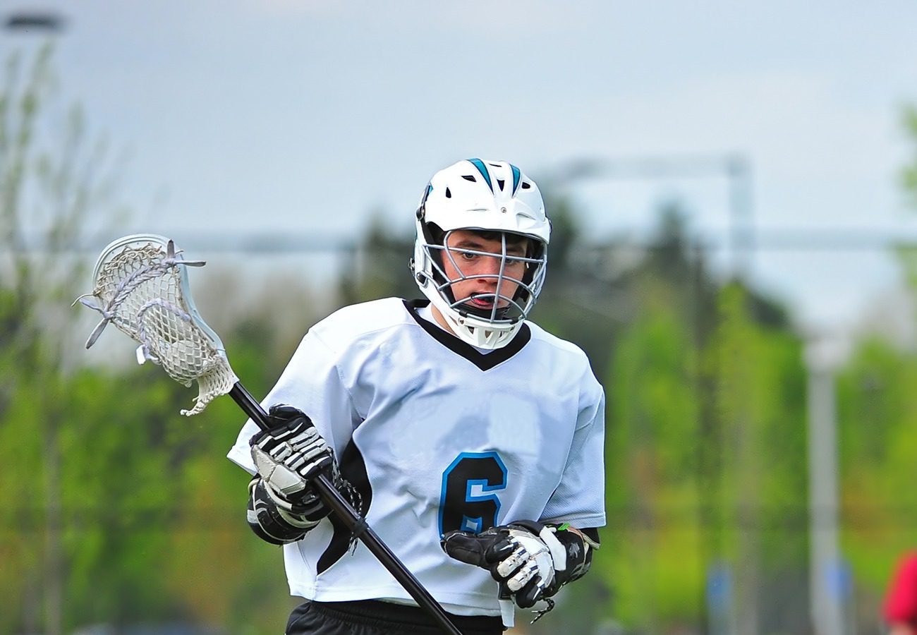 A lacrosse player in a blue jersey and black helmet crouches in an athletic stance on a grassy field, ready for action.
