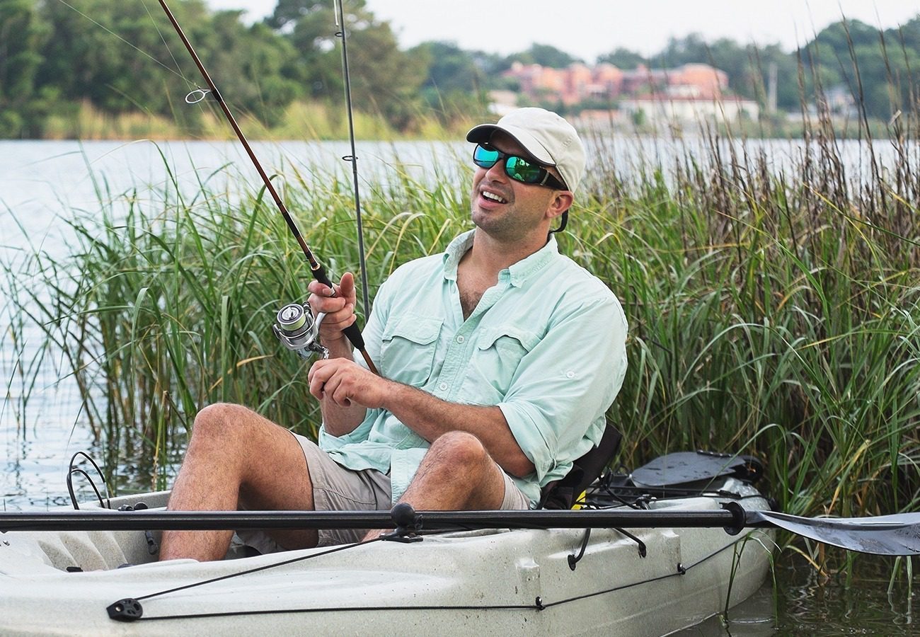 A man wearing sunglasses, a light-colored cap, and a light shirt smiles while fishing from a small boat surrounded by reeds on a lake.