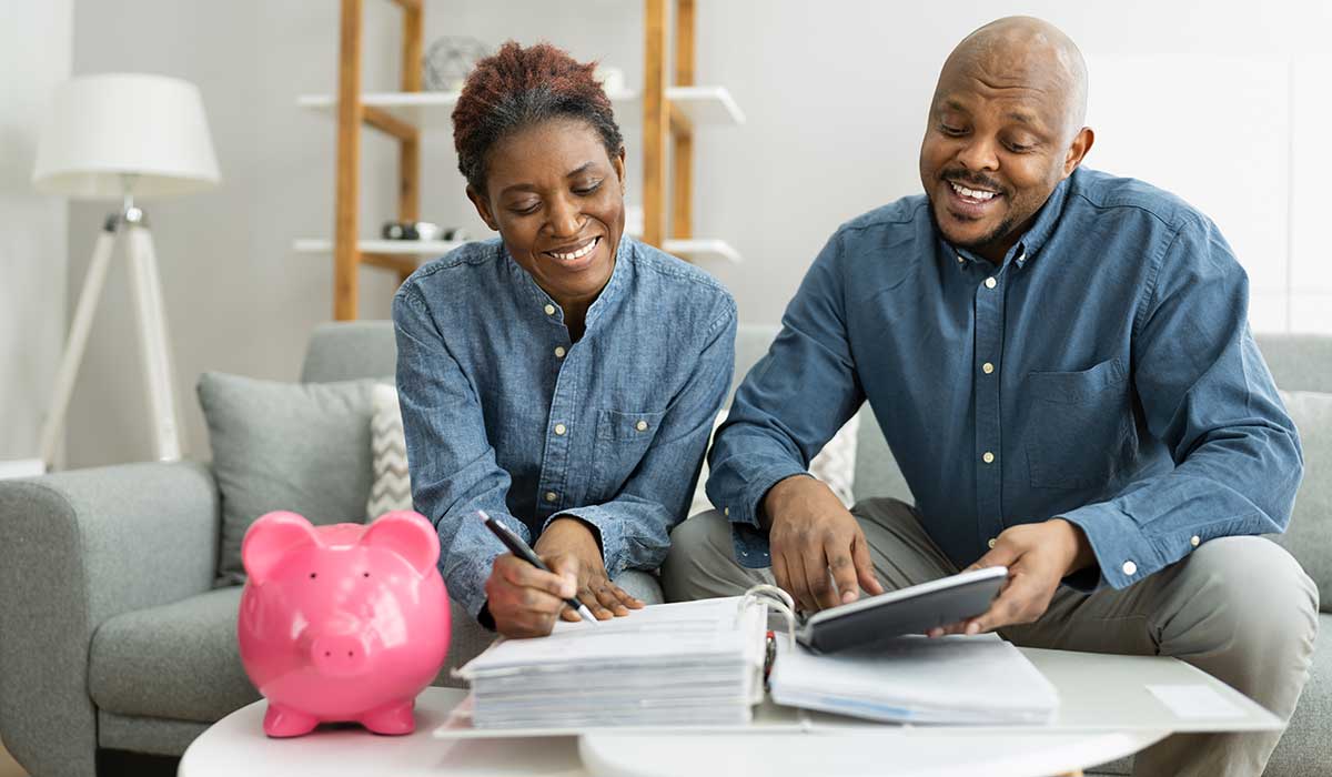 A smiling woman hugs a man from behind as he writes on paper at a kitchen table. Both are happy, surrounded by kitchenware and a laptop, in a cozy, modern kitchen.