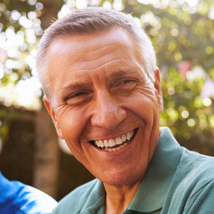 A middle-aged man with short gray hair smiles warmly while standing outdoors in natural sunlight, wearing a green collared shirt. The background is blurred with greenery.