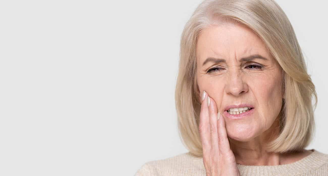 Older woman with short blond hair holds her hand to her cheek and winces in pain, possibly experiencing a toothache or jaw discomfort, against a plain light background.