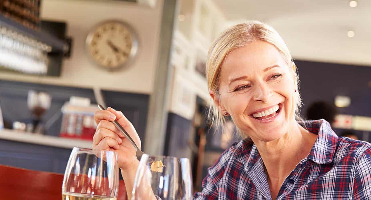 A smiling older woman with blonde hair sits at a table in a restaurant, holding a fork and talking to someone. She wears a plaid shirt and appears happy and engaged. A clock is visible on the wall in the background.