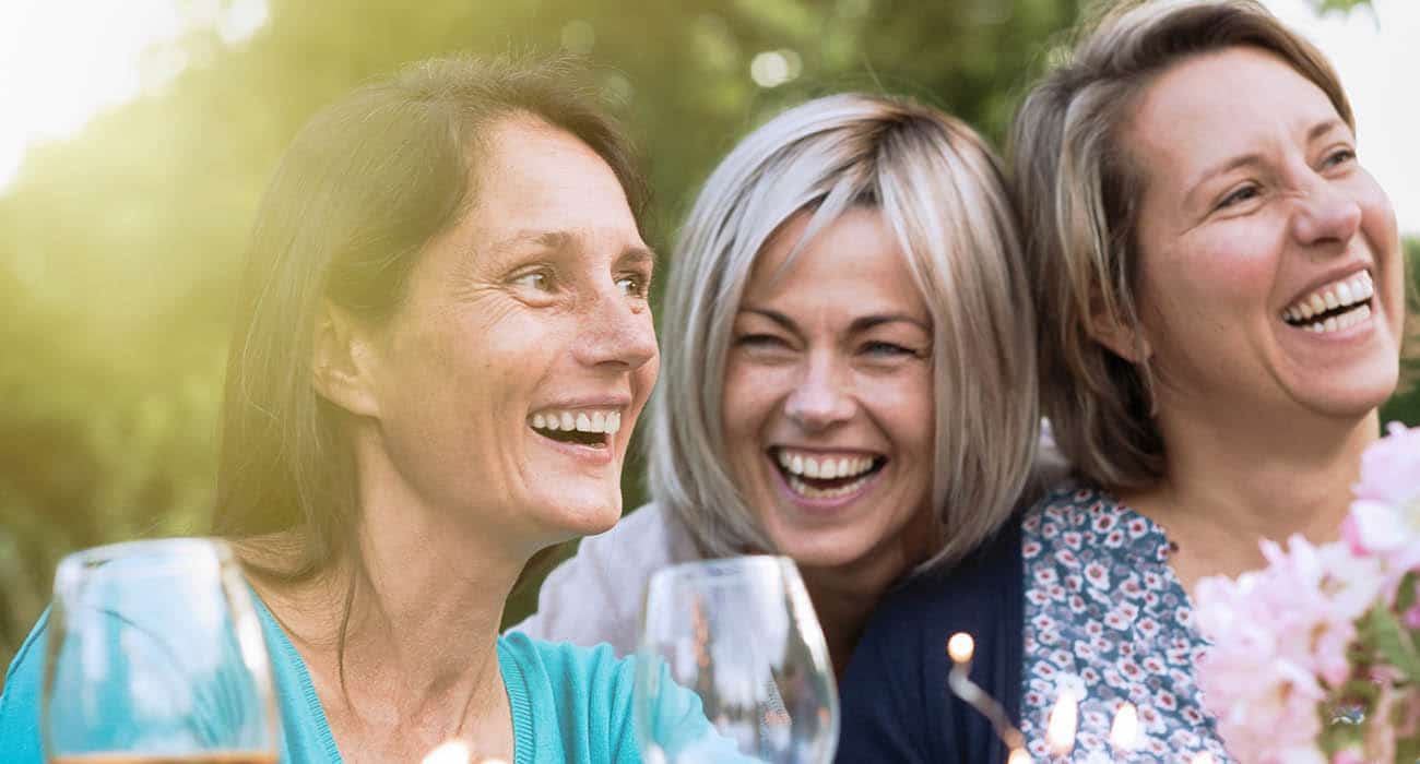A woman and a young girl sit together indoors, smiling and laughing. Sunlight streams through a window behind them, with green potted plants on the windowsill. Both appear happy and relaxed.