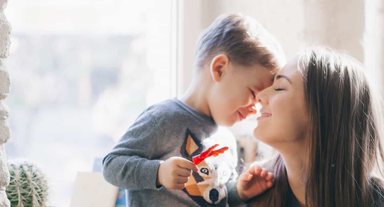 A young child and a woman smile with their faces touching gently in a sunlit room. The child wears a gray shirt with an animal graphic, and both appear joyful, sharing a tender and happy moment together.