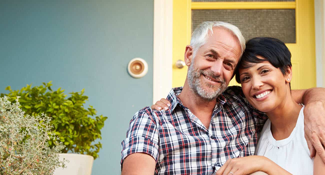 A smiling couple walks outdoors, holding bicycle handlebars. The woman has her arm around the man, and they look at each other happily. They are surrounded by greenery on a sunny day.