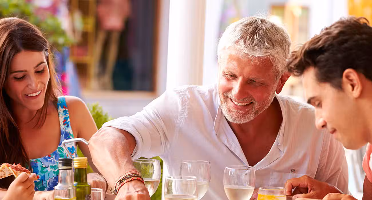 Two men sit outdoors at a table, smiling and enjoying a meal with pizza and drinks. There are several glasses and plates on the table, and other people are partially visible in the background.