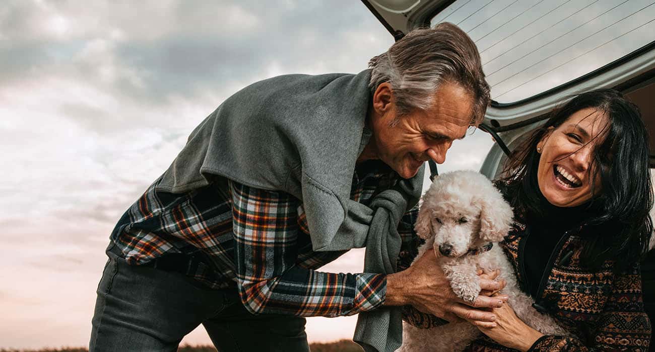 Three men sit close together outdoors, smiling and laughing. The man in the center, wearing a blue shirt, is the focus, while the other two men are partially visible, engaged and joyful. Sunlight filters through trees in the background.