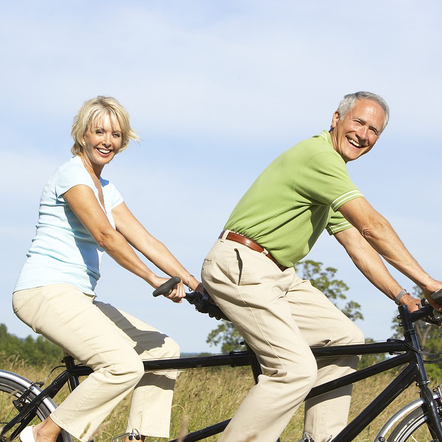 A man holds a laughing young girl outdoors. They are both smiling, enjoying a sunny day together in a green, blurry park. The man has short dark hair and a beard; the girl has long blonde hair and a light shirt.
