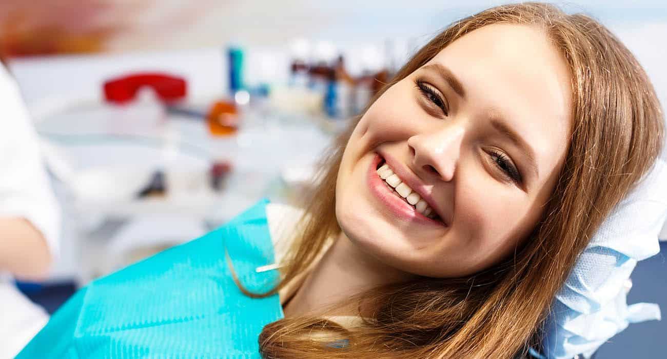 A young woman with long, light brown hair smiles brightly while reclining in a dental chair, wearing a blue dental bib. The background shows blurred dental equipment.